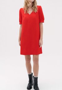 Vestido rojo con escote en V, mangas cortas abullonadas y corte holgado. Lleva botas negras hasta el tobillo. Tejido suave y largo por encima de la rodilla.