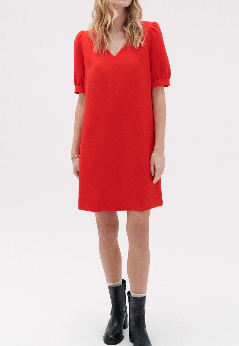 Vestido rojo con escote en V, mangas cortas abullonadas y corte holgado. Lleva botas negras hasta el tobillo. Tejido suave y largo por encima de la rodilla.