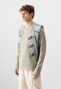 Lys grå vest med et strømlinet design, der har justerbare stropper, en lomme og en blød tekstur, båret over en afdæmpet grøn langærmet skjorte.