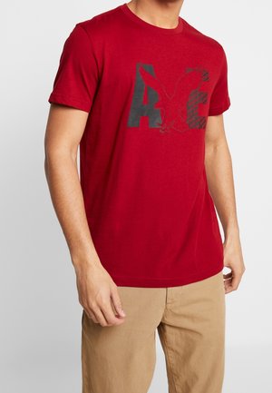 Uomo che indossa una t-shirt rossa a maniche corte con la grafica di un'aquila nera e le lettere A e E, abbinata a pantaloni marroni chiari.