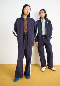 Δύο άτομα φοράνε ταιριαστά σκούρα μπλε αθλητικά κοστούμια με λευκές και μέντα ρίγες. Η γυναίκα έχει ένα τοπ με πορτοκαλί μοτίβο, και και οι δύο φοράνε αθλητικά παπούτσια.