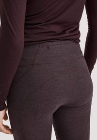 Close-up van een persoon in donkerpaarse, aansluitende leggings en een bijpassend lange mouw top, met gedetailleerde stiksels en een kleine taille-rits.