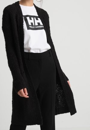 Lange zwarte cardigan gemaakt van textuurgebreid materiaal. Open voorkant ontwerp met een losse pasvorm, gedragen over een wit T-shirt en zwarte broek.