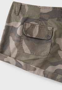 Green and brown camouflage mini skirt with a belt loop and a large front patch pocket with a flap, viewed close up against a white background.