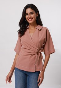 Blusa avvolgente rosa chiaro con maniche corte, colletto, fiocco laterale e dettagli di cucitura bianchi. Indossata con jeans in denim blu.