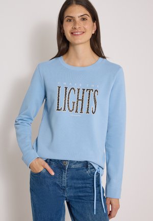 Młoda kobieta o długich ciemnych włosach nosi jasnoniebieską bluzę z tekstem "CHASE THE LIGHTS" w połączeniu z niebieskimi dżinsami, delikatnie się uśmiechając.