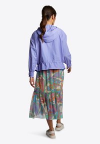 Světle fialová zkrácená bunda s kapucí, spárovaná s barevným vzorovaným maxi šaty, které obsahují odstíny zelené, růžové a oranžové.