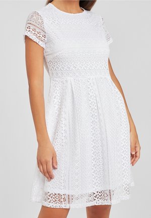 Mujer con vestido blanco de encaje hasta la rodilla, con mangas cortas y escote redondo, de pie sobre un fondo gris liso.
