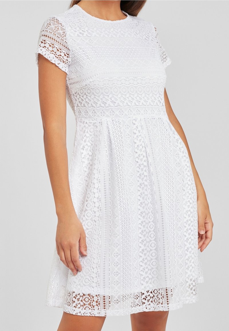 Femme portant une robe blanche en dentelle longueur genou, avec manches courtes et col rond, debout devant un fond gris uni.
