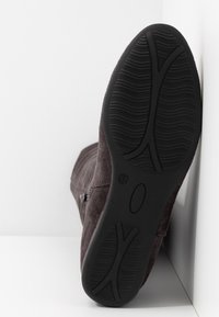Semelle d'une botte en daim brun foncé avec une semelle extérieure en caoutchouc noir texturé, fermeture éclair latérale visible, présentée sur fond blanc.