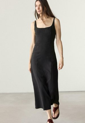 Frau mit langen braunen Haaren, die ein ärmelloses schwarzes Midikleid und burgunderrote Sandalen trägt, vor einem einfarbigen hellen Hintergrund gehend.