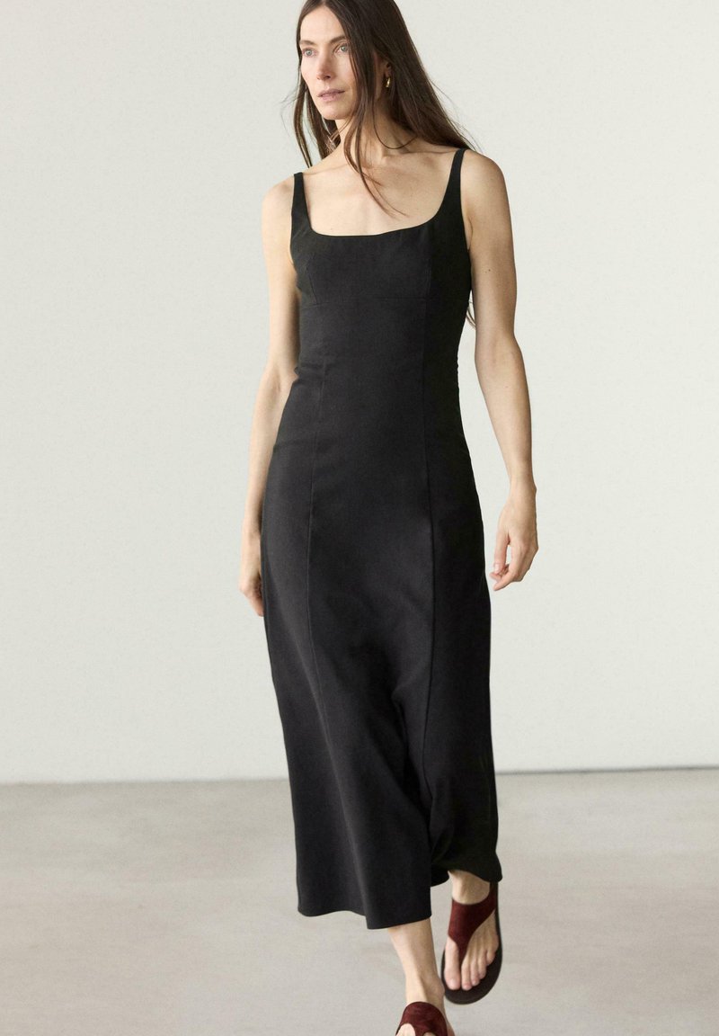 Kobieta z długimi brązowymi włosami, ubrana w czarną sukienkę midi bez rękawów i burgundowe sandały, idąca na tle jasnego, jednolitego tła.