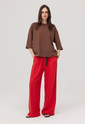 Kobieta stoi z rękami w kieszeniach, ubrana w luźną brązową bluzkę z krótkim rękawem, jasnoczerwone szerokie spodnie i czarne sandały z odkrytymi palcami.