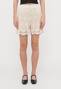 Lace shorts in light beige with floral pattern, scalloped hem, and elastic waistband, paired with black patent shoes.