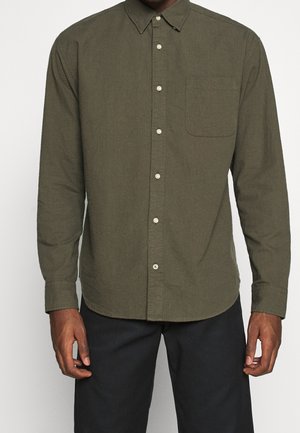 Mężczyzna w oliwkowej długiej koszuli na guziki z kieszenią na piersi i czarnych spodniach, stojący na białym tle.