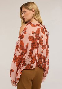 Blusa rosa trasparente con bordi arricciati e spalle scoperte, caratterizzata da un grande motivo floreale rosso; tessuto leggero e vestibilità ampia.
