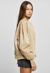 Giubbotto bomber beige in tessuto lucido, dalla vestibilità oversize, con polsini e colletto a costine. Abbinato a pantaloni corti scuri, che mostrano una silhouette rilassata.
