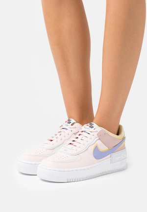 Osoba nosząca różowe sneakersy Nike Air Force 1 z fioletowymi i żółtymi akcentami, stojąca na białej powierzchni.