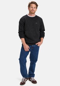 Sudadera negra con ribete blanco en el cuello, que presenta un pequeño logo. Combinada con jeans de mezclilla azul y zapatillas marrones con rayas blancas.