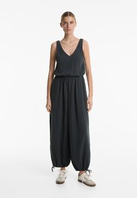 Črn kombinezon z V-izrezom, brez rokavov, z zbranim pasom in zoženimi gležnji. Nosi se ga s svetlo bež supergami. Gladka tekstura.