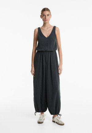 Combinaison noire avec un décolleté en V, sans manches, taille froncée et chevilles fuselées. Portée avec des baskets beiges. Texture lisse.