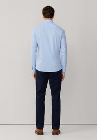 Lichtblauw blouse met een subtiel patroon, lange mouwen en een gebogen zoom, gecombineerd met donkerblauwe broek en bruine schoenen.