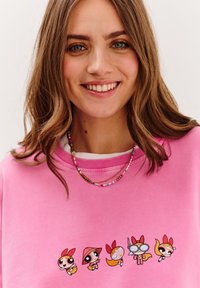 Różowy sweatshirt z pięcioma animowanymi postaciami wyszytymi na przodzie. Odzież ma klasyczny okrągły dekolt i ściągacze przy rękawach.