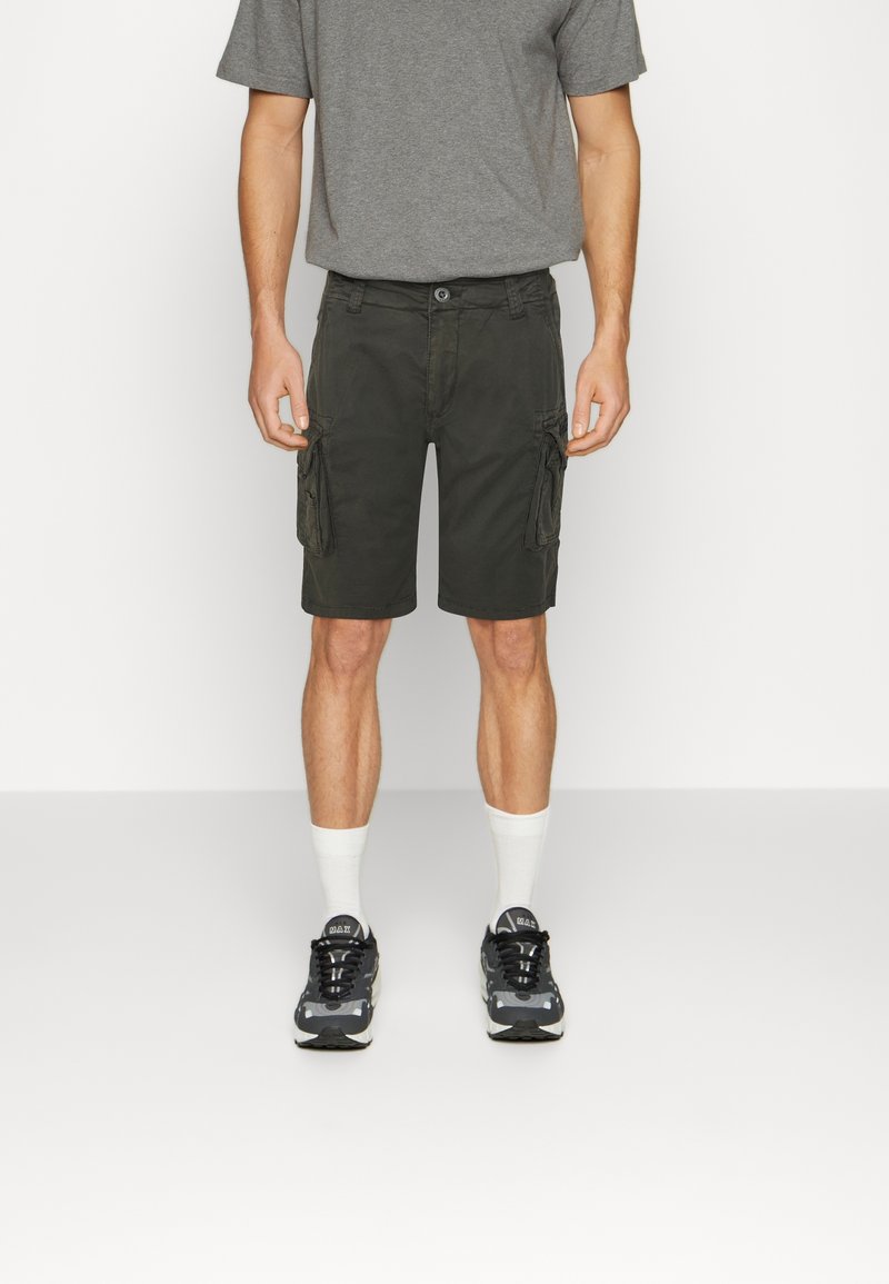 Homme portant un short cargo foncé, un t-shirt gris, des chaussettes blanches et des baskets noires, debout devant un fond blanc uni.