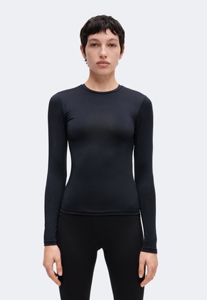Persona joven con cabello corto y oscuro que lleva una camiseta negra ajustada de manga larga y pantalones negros, de pie contra un fondo claro y liso.
