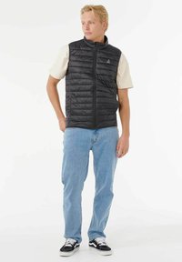 Mand iført sort quiltet vest over hvid kortærmet skjorte, lyseblå jeans og sort-hvide sneakers, stående mod hvid baggrund.