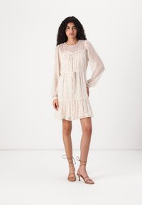 Robe beige clair avec un ourlet à volants, des manches longues et des pois brodés. Dotée d'une taille à cordon pour un ajustement réglable.