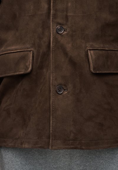 Μπροστινή όψη σκούρου καφέ σατέν μπουφάν με δύο τσέπες με καπάκι και δύο ορατά κουμπιά πάνω από γκρι παντελόνι από ύφασμα.