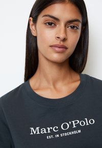Mørkegrå T-shirt med rund hals, prydet med hvid tekst "Marc O'Polo EST. IN STOCKHOLM." Glat stof, afslappet design.