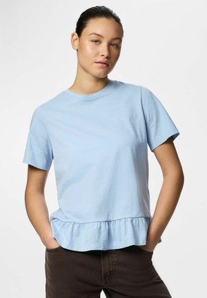 Ung kvinne med håret bundet bak, iført en lyseblå topp med korte ermer og rysjekant, samt mørke bukser, stående med hendene i lomma.