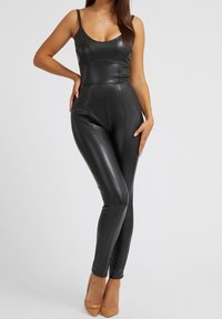 Frau in einem figurbetonten schwarzen, ärmellosen Catsuit aus Kunstleder und beigen spitzen High Heels, die mit einer Hand auf der Hüfte steht.
