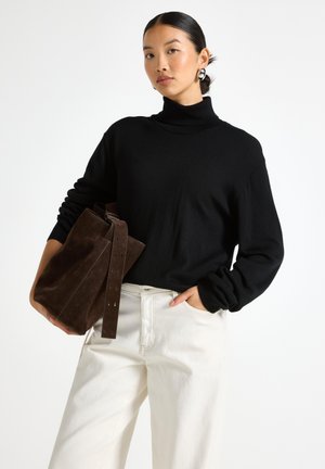 Maglione a collo alto nero in maglia con maniche lunghe abbinato a pantaloni a gamba larga crema, tiene una borsa in suede marrone scuro con dettagli sulla tracolla.