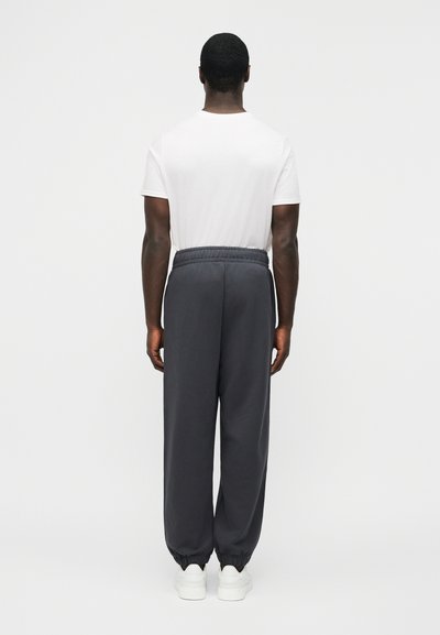 Άνδρας με σκουρόχρωμο δέρμα που φοράει λευκό μπλουζάκι, φαρδιά σκούρα γκρι φόρμα, και λευκά αθλητικά παπούτσια, στέκεται αντικρίζοντας έναν απλό φωτεινό φόντο.