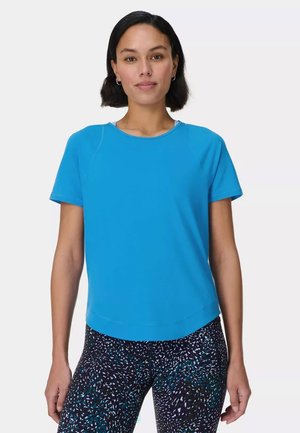 Vrouw met kort donker haar, gekleed in een felblauw shirt met korte mouwen en zwarte legging met blauw-wit gespikkeld patroon, staand tegen een witte achtergrond.