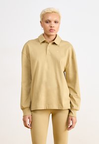 Polo a maniche lunghe color khaki con bottoni, fessure laterali e logo Nike sul petto, abbinato a leggins coordinati.