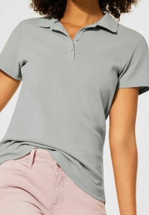 Frau trägt ein hellgraues, kurzärmeliges Poloshirt, das in eine hellrosa Hose gesteckt ist, vor weißem Hintergrund.