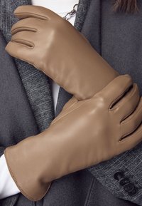 Guanti in pelle marrone dalla texture liscia, con dita allungate e polsino aderente. Indossati sopra un cappotto grigio con tessuto texturizzato.