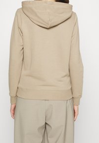 Beige mikina s kapucí na šňůrku, jemná textura, žebrované manžety a lem, zobrazená ze zadní strany s uvolněným střihem.