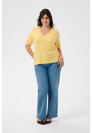 Femme debout avec un léger sourire, portant un pull jaune à manches courtes, un jean bleu et des chaussures marron pointues sur fond blanc.