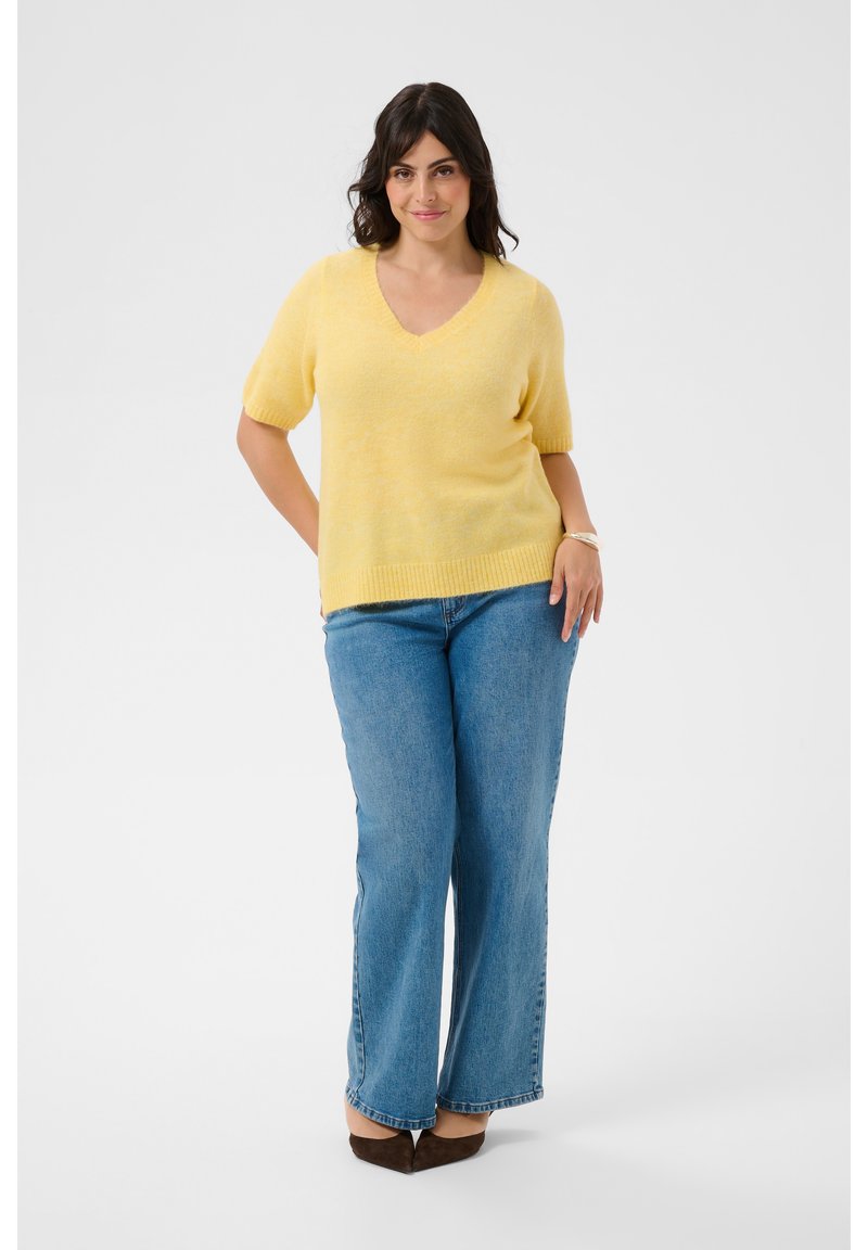 Femme debout avec un léger sourire, portant un pull jaune à manches courtes, un jean bleu et des chaussures marron pointues sur fond blanc.