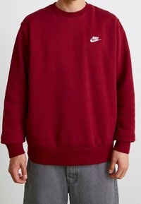 En person klädd i en enkel röd Nike-sweatshirt med långa ärmar och grå jeans, stående mot en ljus, enfärgad bakgrund.