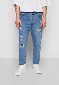 Světle modré džínové kalhoty s roztrhaným designem a zužujícím se střihem. Mají opotřebované části a třásně na lemu, doplněné bílými teniskami.