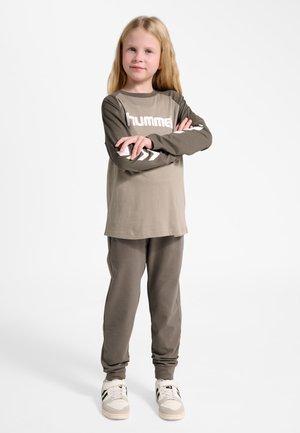 Junges Mädchen mit langen blonden Haaren, das einen beigen und olivfarbenen Hummel-Sweatshirt und passende Hose trägt und mit verschränkten Armen steht.