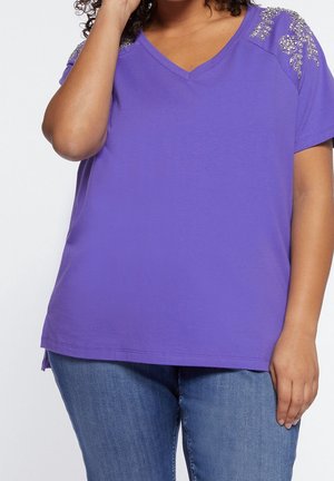 Camiseta morada de cuello en V con mangas cortas, adornada con acentos plateados en los hombros, hecha de tela suave, combinada con jeans de mezclilla.