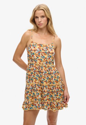 Mujer con vestido sin mangas amarillo con flores azules y rojas, de pie con un brazo doblado y expresión neutral.