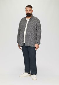 Une chemise à carreaux noire et blanche, portée par-dessus un t-shirt clair, associée à un pantalon foncé et des baskets blanches, se tenant contre un fond neutre.